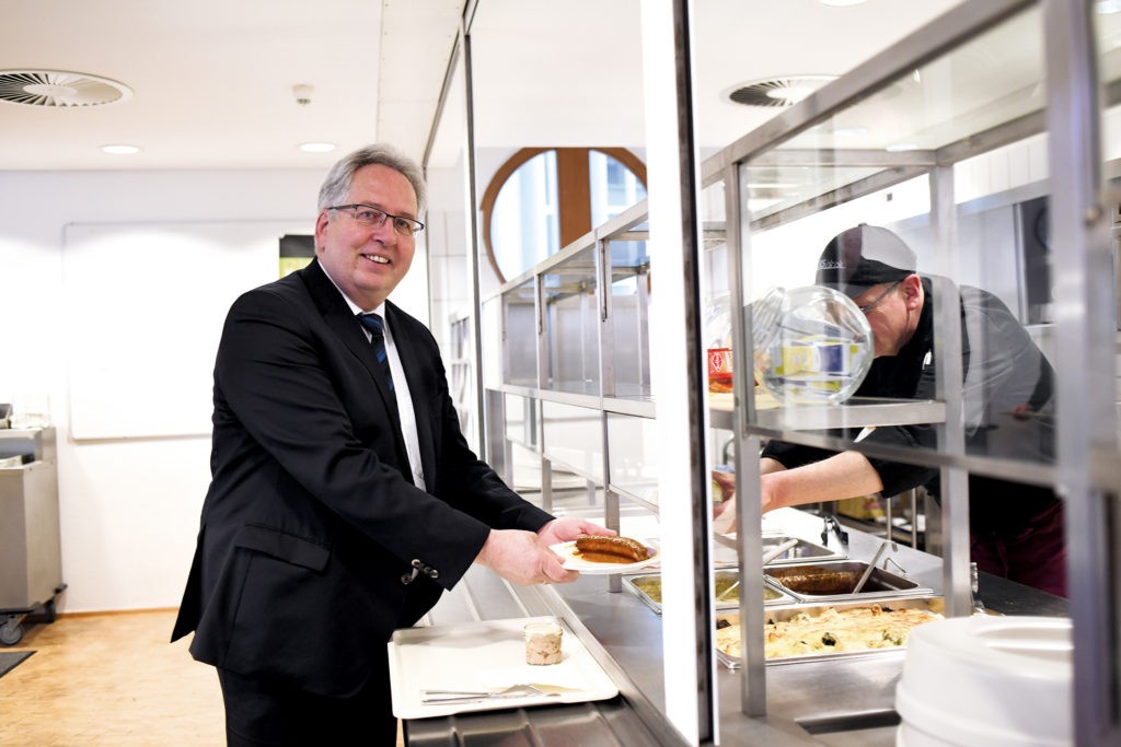 Jürgen Müller ist in Anzug und Schlips gekleidet; er nimmt an der Essenstheke der Kantine gerade einen Teller mit Currywurst von einem Küchenmitarbeiter entgegen und lächelt dabei in die Kamera.
