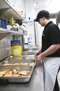 Ein Mitarbeiter steht in Arbeitskleidung in der Großküche; man sieht ihn von der Seite. Er schiebt gerade Edelstahl-Behälter nebeneinander, in denen geröstete Brotscheiben liegen.