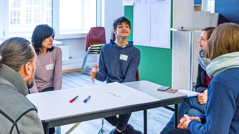 Blick in eine Workshop-Runde, in der mehrere Menschen um einen Tisch herum sitzen. Einer davon hat eine körperliche Behinderung und erklärt auf dem Foto gerade etwas.