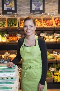 Mercedes Hamann steht mit grüner Schürze vor dem Obst- und Gemüseregal im Hofladen des Hofguts Schloss Hamborn und lächelt in die Kamera.
