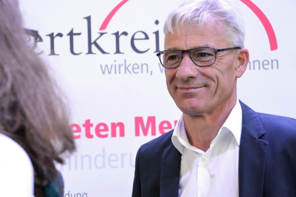 Ulrich Rötgers, im Hintergrund ein weißes Plakat mit der teilweise lesbaren Aufschrift "wertkreis".