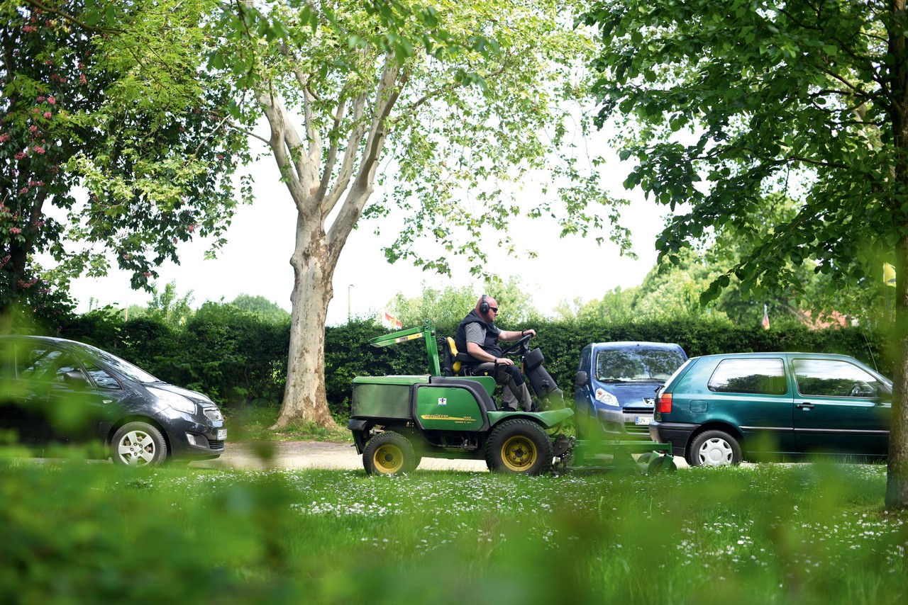 Ein Mitarbeiter der Grünbau fährt auf einem Mähtraktor an der Straße entlang, im Vordergrund eine grüne Wiese und Büsche.