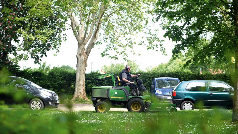Ein Mitarbeiter der Grünbau fährt auf einem Mähtraktor an der Straße entlang, im Vordergrund eine grüne Wiese und Büsche.