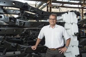 Der Geschäftsführer von Teuto InServ steht lächelnd in einer Lagerhalle vor einem Regal voller großer Autobauteile.