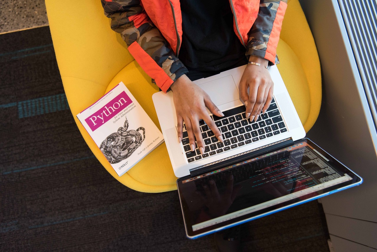 Die Hände einer Frau auf der Tastatur eines Laptops von oben, neben ihr liegt ein Buch zu einer Programmiersprache.