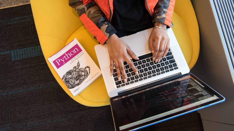 Die Hände einer Frau auf der Tastatur eines Laptops von oben, neben ihr liegt ein Buch zu einer Programmiersprache.
