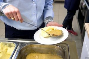 Eine Servicekraft schöpft das Mittagsmenü aus den Warmhaltewannen auf einen Teller. Es sind nur die Hände und der Teller zu sehen.