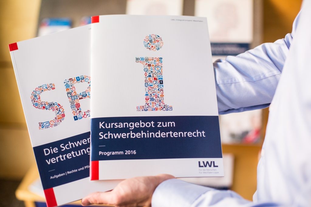 Ein Mitarbeiter des LWL-Integrationsamtes zeigt zwei Hefte aus der Infothek, unter anderem das Kursangebot des Amtes zum Schwerbehindertenrecht.