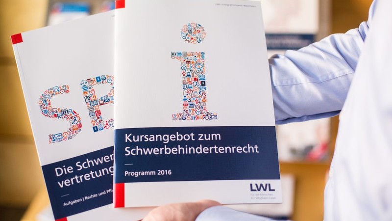 Ein Mitarbeiter des LWL-Integrationsamtes zeigt zwei Hefte aus der Infothek, unter anderem das Kursangebot des Amtes zum Schwerbehindertenrecht.
