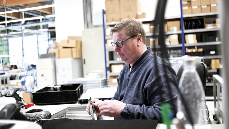 Ein Mitarbeiter der AfB sitzt in der Lagerhalle des Unternehmens an seinem Werktisch bearbeitet und konzentriert ein Computer-Bauteil, das er in der Hand hält.