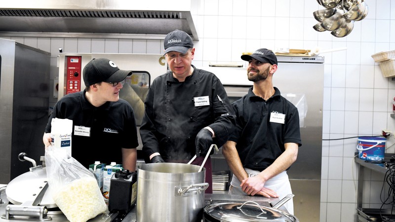 enedikt Minnig, Frank Kölling und Aqid Ibrahim (von links) stehen in Arbeitskleidung in der Großküche des Unternehmens hinter einem großen Kochtopf, Frank Kölling rührt, die drei sprechen lachend miteinander.