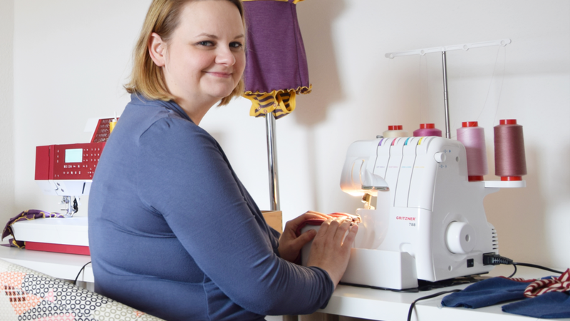 Sandra Brunner sitzt an ihrer Nähmaschine und schaut über die Schulter zurück in die Kamera.