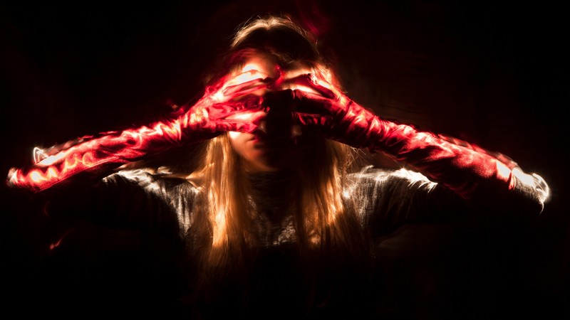 Eine Frau vor tiefschwarzem Hintergrund mit langen Haaren und roten Handschuhen hält sich die Augen zu. Die Handschuhe und die Haare leuchten.