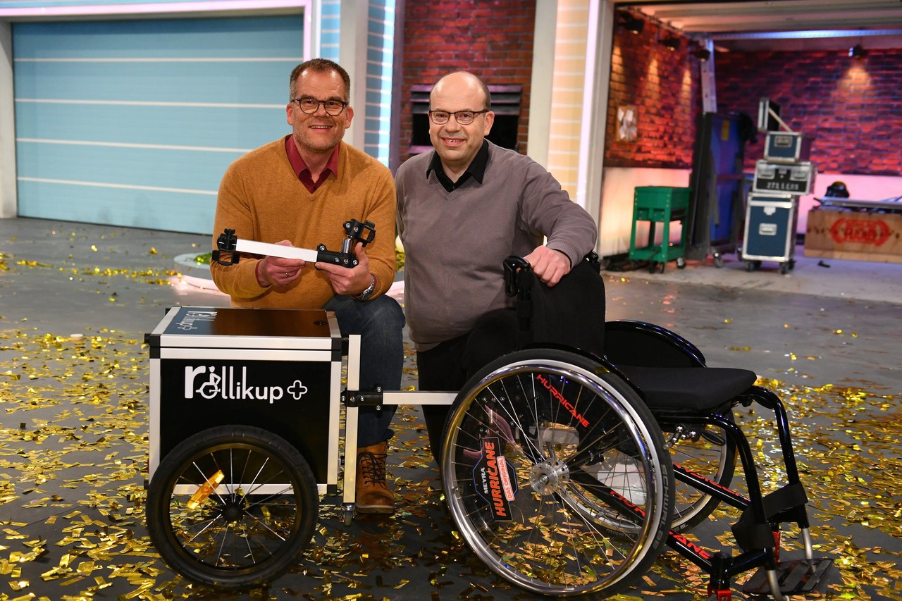 Andreas Neitzel (links), Eduard Wiebe (rechts) posieren mit einem Rollstuhl samt Rollikup und Anhänger nach der Show auf der Bühne.