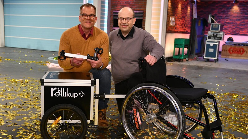 Andreas Neitzel (links), Eduard Wiebe (rechts) posieren mit einem Rollstuhl samt Rollikup und Anhänger nach der Show auf der Bühne.