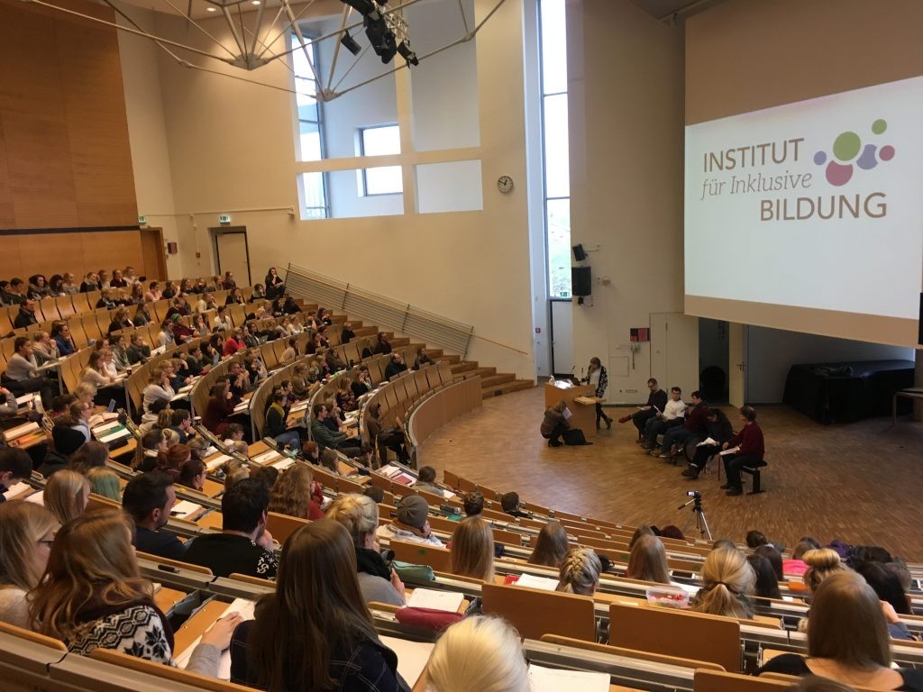 Blick in einen vollen Hörsaal der Europa-Universität in Flensburg, vorne vier Bildungsfachkräfte des Instituts für Inklusive Bildung, die die Vorlesung halten.