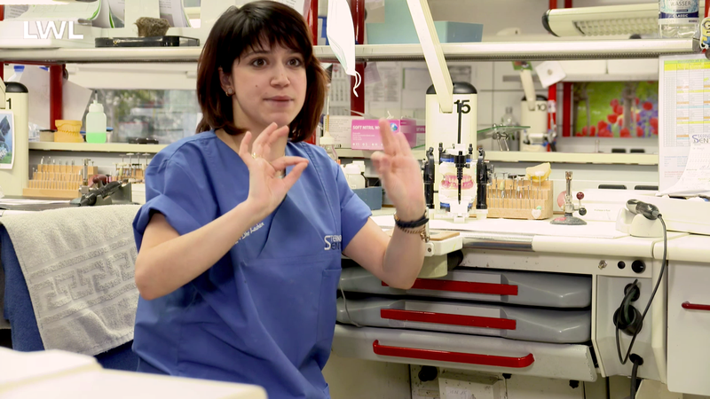 Eine gehörlose Zahntechnikerin des Dentallabors erklärt in Gebärdensprache etwas in die Kamera.