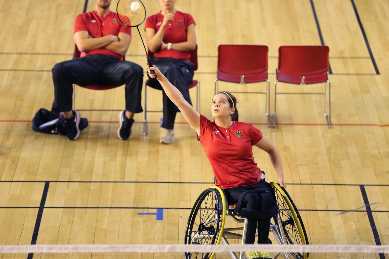 Valeska Knoblauch spielt in rotem Turnier-Trikot Para-Badminton in einer Sporthalle. Sie ist von vorne bei der Ballannahme zu sehen.