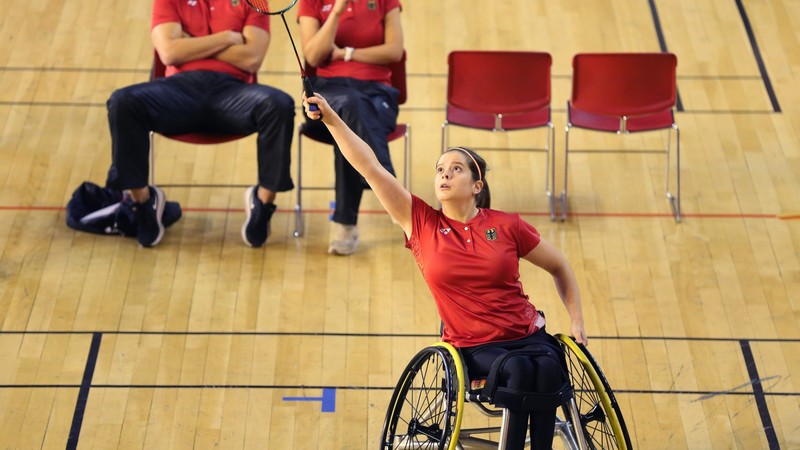 Valeska Knoblauch spielt in rotem Turnier-Trikot Para-Badminton in einer Sporthalle. Sie ist von vorne bei der Ballannahme zu sehen.
