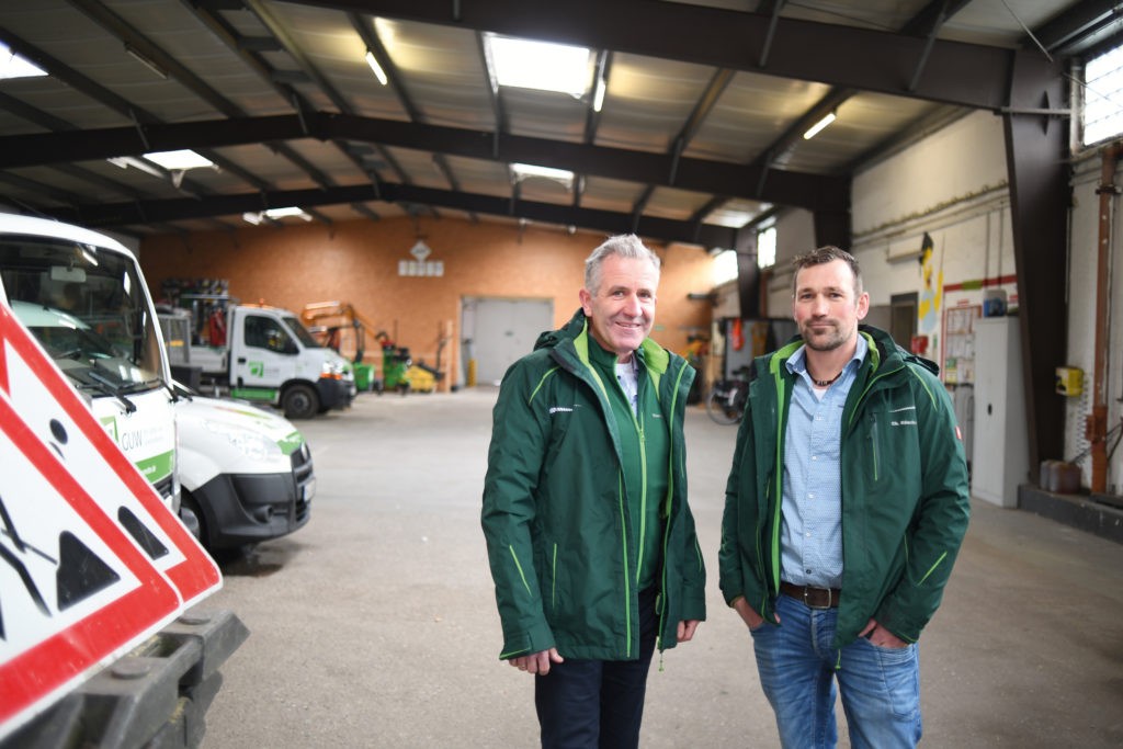 Thomas Pahls (links) und Christian Rüschoff stehen nebeneinander in grünen Arbeitsjacken in einer große Halle mit Autos und Verkehrsschildern und schauen in die Kamera.