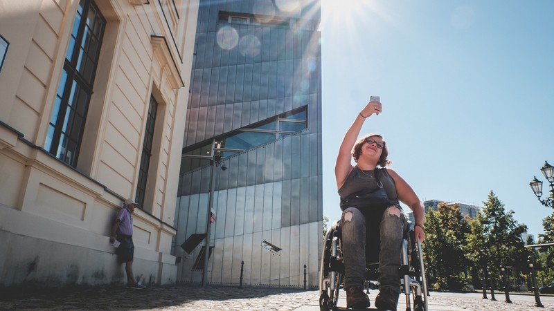 Eine Frau mit Rollstuhl reißt die Faust in die Luft, während sie an einem Gebäude vorbeifährt.