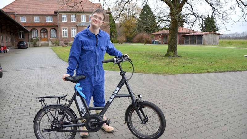 Tobias Koddebusch steht mit seinem Fahrrad auf dem Hof vor dem Stall und lächelt in die Kamera. Er trägt einen blauen Overall.