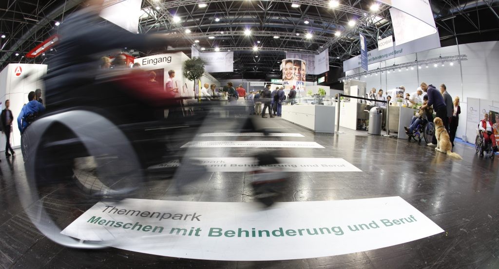 Bewegungsunscharfes Motiv eines Menschen im Rollstuhl, der über die Bodenbeschriftung "Menschen mit Behinderung im Beruf" auf der Rehacare-Messe fährt.