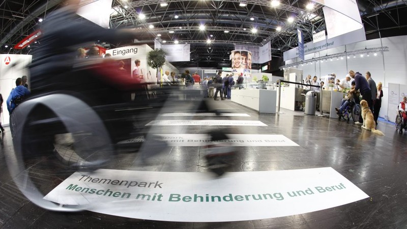 Bewegungsunscharfes Motiv eines Menschen im Rollstuhl, der über die Bodenbeschriftung "Menschen mit Behinderung im Beruf" auf der Rehacare-Messe fährt.