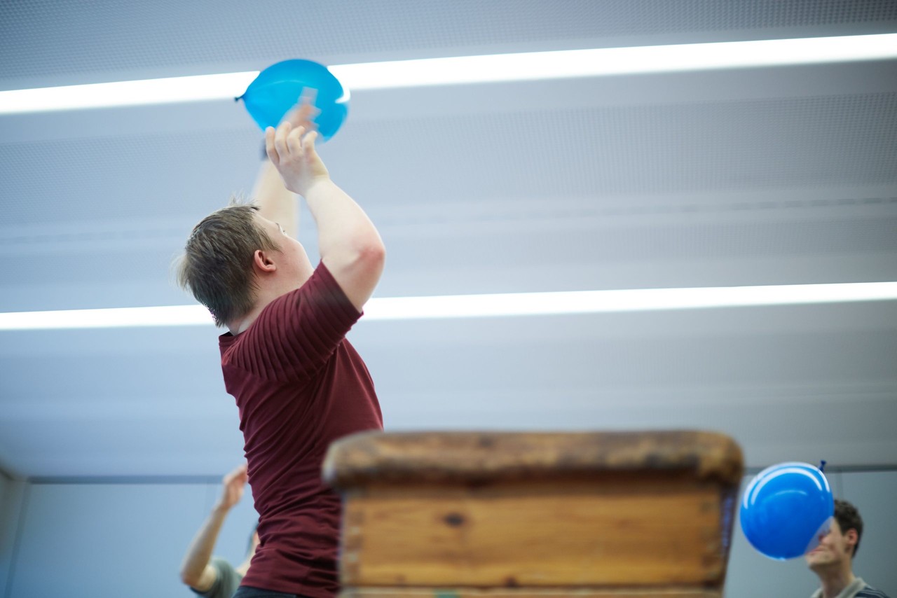 Ein Junge in Sportklamotten in einer Turnhalle mit einem blauen Luftballon in der Hand, fotografiert im Sprung nach oben.