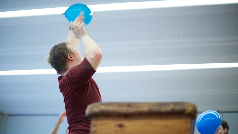 Ein Junge in Sportklamotten in einer Turnhalle mit einem blauen Luftballon in der Hand, fotografiert im Sprung nach oben.