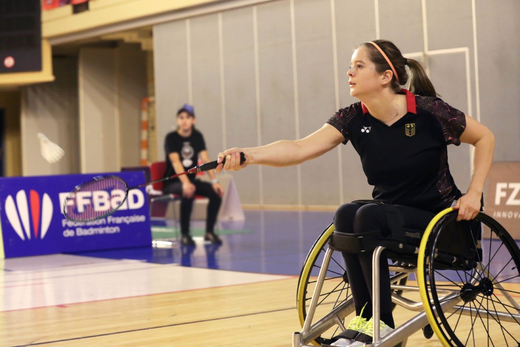 Valeska Knoblauch spielt in schwarzem Trikot Para-Badminton in einer Sporthalle. Sie ist von der Seite bei der Ballannahme zu sehen, im Hintergrund unscharf ein Balljunge am Spielfeldrand.