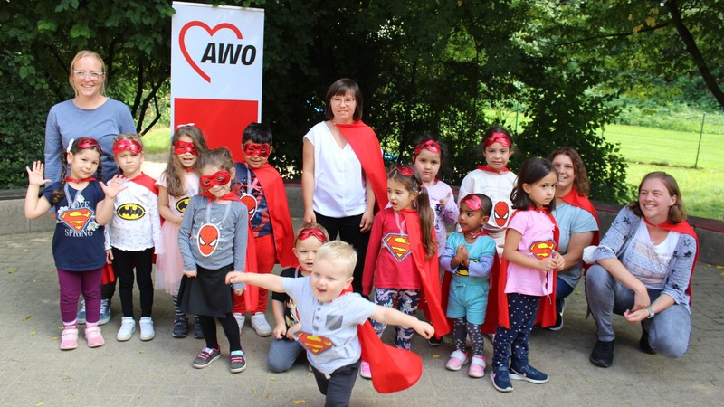 Kinder mit Superhelden-Kostümen haben sich draußen zum Gruppenfoto aufgestellt, vorne tanzt ein Kind mit rotem Umhang aus der Reihe