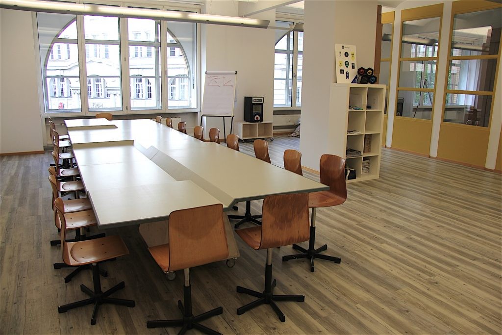 Ein großer, leerer Tisch, dessen Tischplatte verschiedene Höhen hat. Er steht in den Räumen des Coworking-Spaces in Berlin. Drumherum stehen ordentlich viele Stühle.