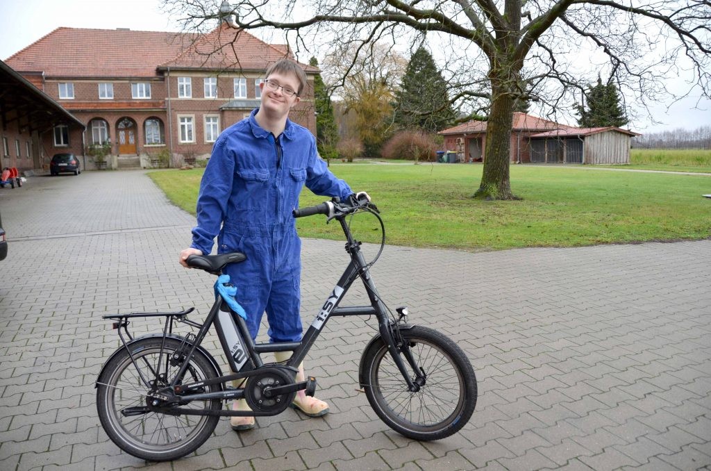 Tobias Koddebusch steht mit seinem Fahrrad auf dem Hof vor dem Stall und lächelt in die Kamera. Er trägt einen blauen Overall.