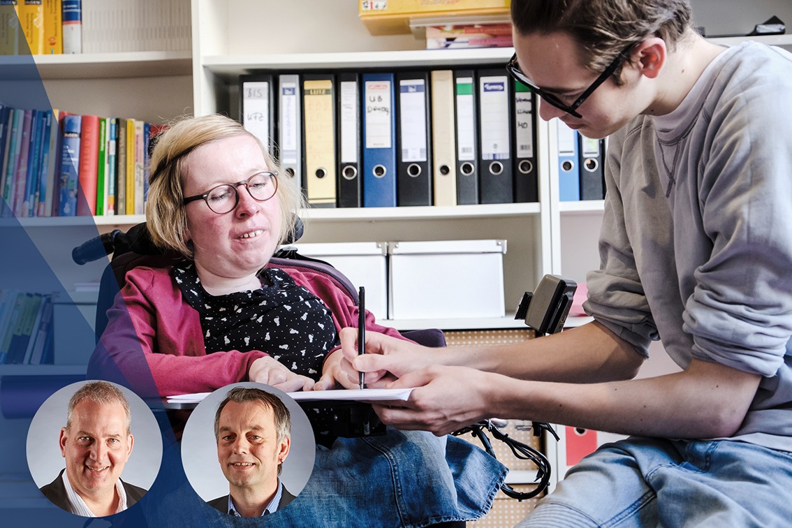 Eine Frau mit Rollstuhl und ein Auszubildender mit Lernbehinderung sitzen zusammen über einem Formular und tragen etwas ein.