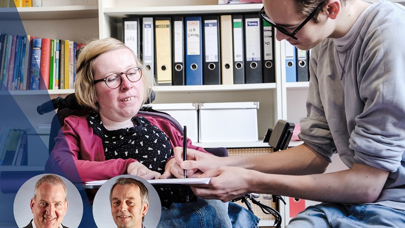 Eine Frau mit Rollstuhl und ein Auszubildender mit Lernbehinderung sitzen zusammen über einem Formular und tragen etwas ein.