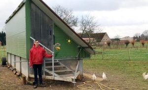 Ein Mitarbeiter des Erlebnis-Bauernhofs steht auf einer Wiese vor einem mobilen Hühnerstall und schaut in die Kamera.