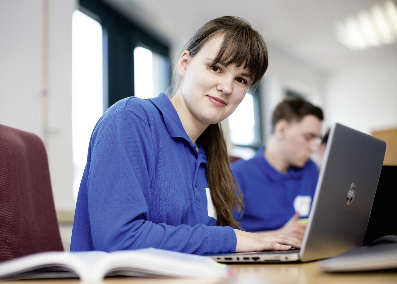 Eine junge Frau sitzt an einem Laptop und schaut in die Kamera.