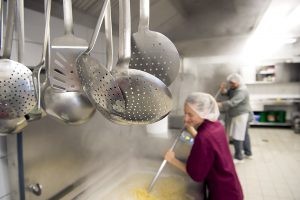Im Vordergrund Küchengeschirr, dahiner eine Frau mit Haube, die mit einem großen Löffel in einer riesigen Kochwanne rührt.