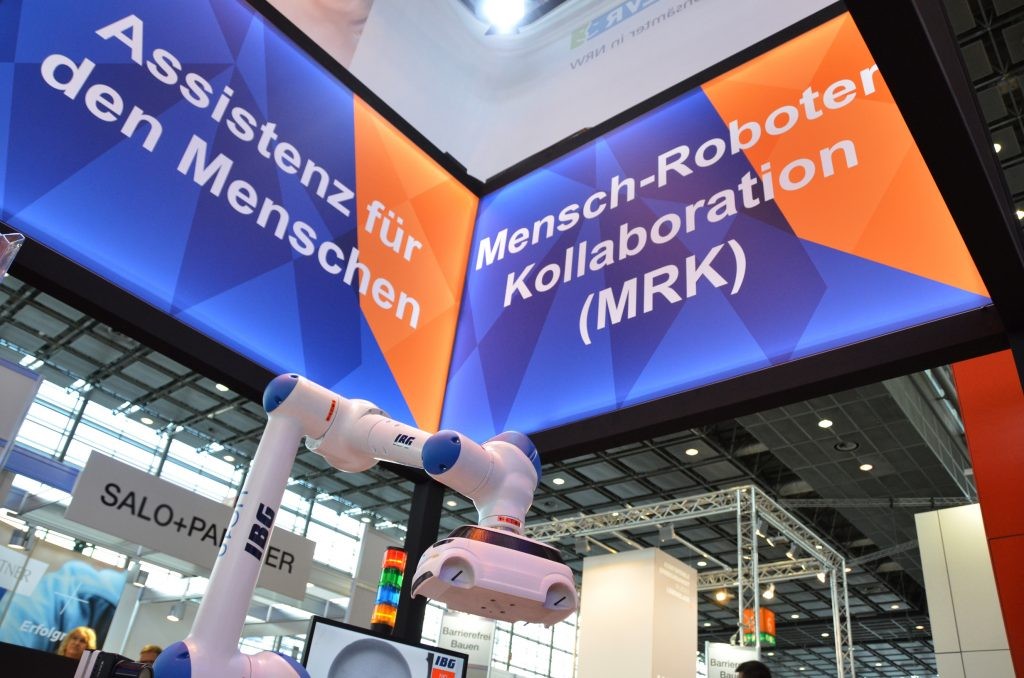 Der IBG-Roboter im Vordergrund unter zwei großen Schildern: auf dem linken Schild steht "Assistenz für den Menschen", auf dem rechten "Mensch-Roboter-Kollaboration (MRK)".