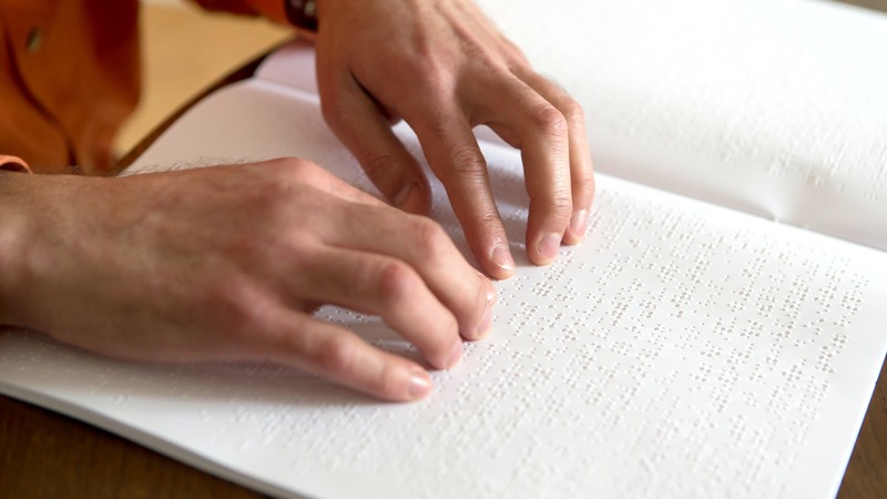Auf dem Foto sind zwei Hände zu sehen, die ein Heft mit Brailleschrift abtasten.