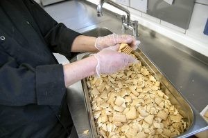 Mitarbeiter Rolf Adämmer zerkleinert mit Handschuhen Butterkekse über einer großen Wanne, die schon fast voll ist. Es sind nur seine Hände und die Wanne zu sehen.