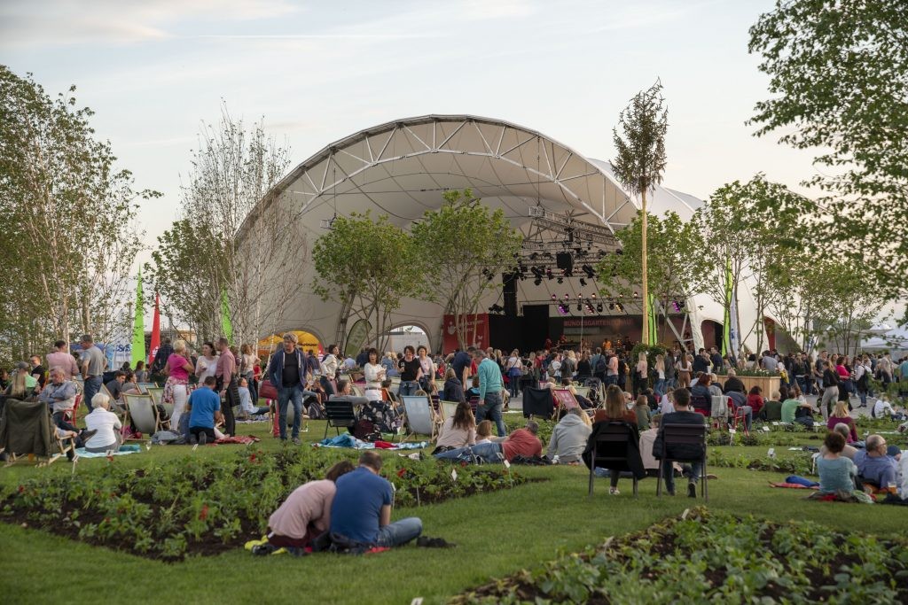 Bei gutem Wetter sitzen und stehen Menschen auf einer Wiese vor einer überdachten Bühne. Foto: BUGA Heilbronn 2019 GmbH