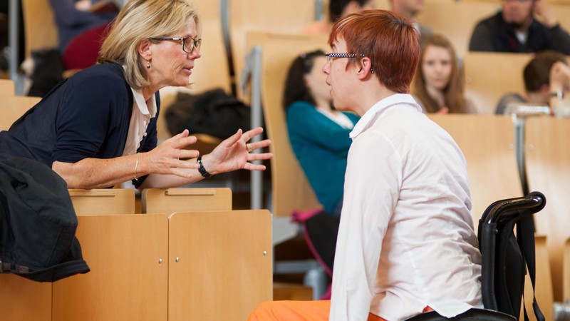 Eine Professorin tauscht sich am Podium in einem vollen Vorlesungssaal mit einer Bildungsfachkraft im Rollstuhl aus.