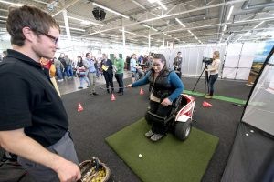 Bei einer Mitmachaktion hat sich eine Besucherin in einen flexiblen Rollstuhl gurten lassen, mit dem sie ohne Beinkraft "aufstehen" und Golf spielen kann.