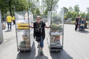 Katharina Wodrich zieht mit der linken und der rechten Hand je ein Rollregal mit Arbeitmaterial über einen Hof mit sich und geht dabei auf die Kamera zu.
