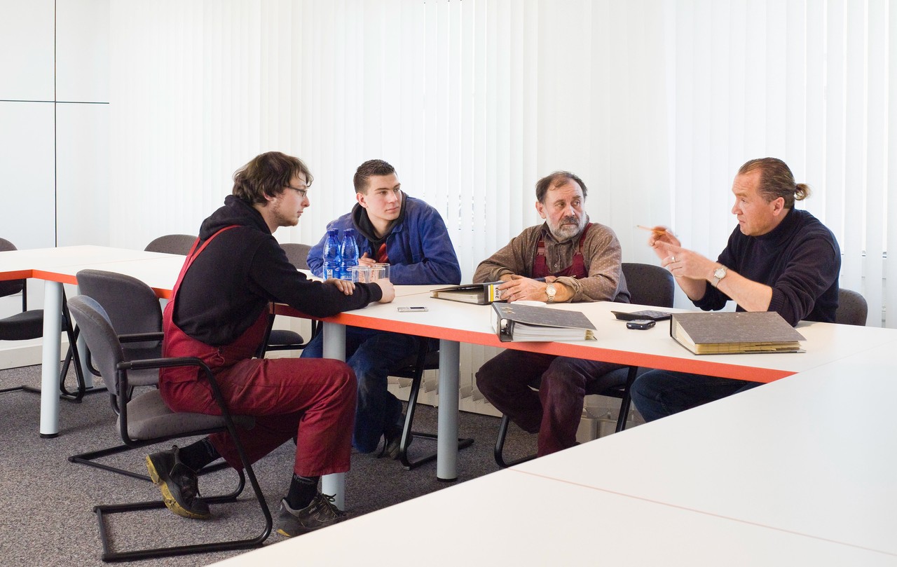 Eine Gruppe Männer in unterschiedlicher Berufskleidung sitzt in einem Tagungssaal an einem Tisch zusammen und unterhält sich.