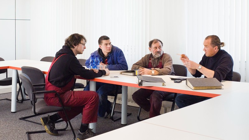 Eine Gruppe Männer in unterschiedlicher Berufskleidung sitzt in einem Tagungssaal an einem Tisch zusammen und unterhält sich.