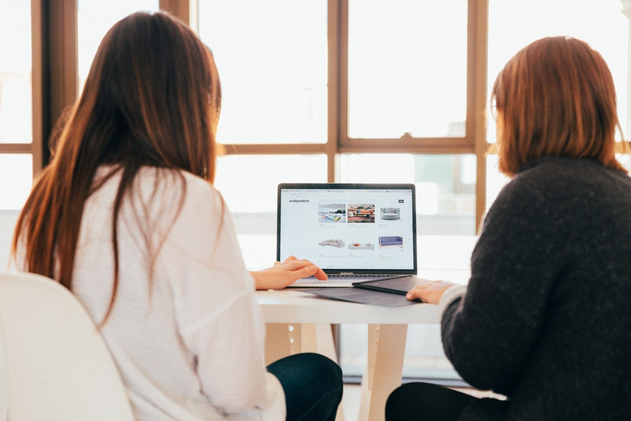 Zwei Frauen sitzen in einem hellen Raum an einem Schreibtisch und schauen gemeinsam auf einen Laptop, auf dem eine Website aufgerufen ist. Beide sind nur von hinten zu sehen, dazwischen der Laptop..