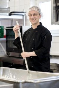 Rolf Adämmer rührt mit einem großen Schneebesen in einer Wanne voller Joghurt und lächelt in die Kamera.